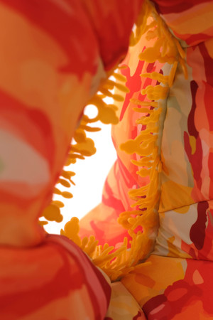 A kinetic sculpture that is made out of soft materials. It extends from floor to the ceiling. It is orange at the base with pink m red and white stripes and orange polka dots. It has a tear drop shape on the lower part where it is orange and a hole in the middle of the tear drop. The hole has little bubble shapes details around it.They look like beads. Midway through the sculpture oranges fades to light green and the shape changes. The tear drop shape is attached on top onto a tubular structure with green  dangling tentacle like pieces. As these pieces move, some orange colors appears in between them where they are attached to the tubular shape. The green pieces move up and down they are light and see through with orange lines and polka dots on them.