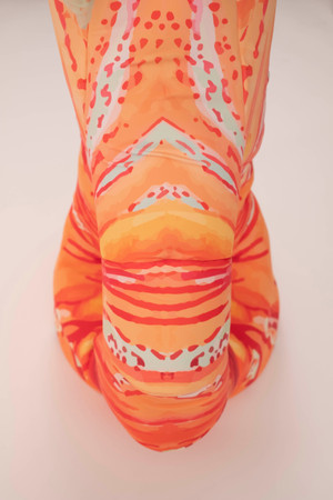 A kinetic sculpture that is made out of soft materials. It extends from floor to the ceiling. It is orange at the base with pink m red and white stripes and orange polka dots. It has a tear drop shape on the lower part where it is orange and a hole in the middle of the tear drop. The hole has little bubble shapes details around it.They look like beads. Midway through the sculpture oranges fades to light green and the shape changes. The tear drop shape is attached on top onto a tubular structure with green  dangling tentacle like pieces. As these pieces move, some orange colors appears in between them where they are attached to the tubular shape. The green pieces move up and down they are light and see through with orange lines and polka dots on them.